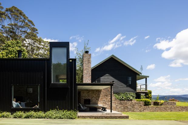 Exterior view of a contemporary dark-clad residential home with a steep gabled roof, projecting upper-level window, brick chimney and brick terrace walls. The house opens onto a paved patio and lawn, with distant hills visible beyond the property.