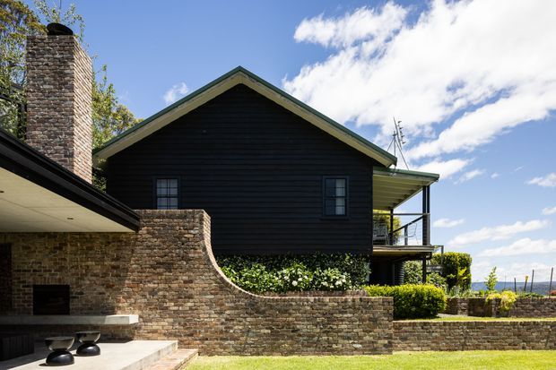 Exterior view of a dark timber-clad gable-roof house with prominent rough stone masonry chimney and curved stone boundary wall. A covered outdoor entertaining area and balcony are visible, with landscaped hedges and lawn in the foreground and open landscape beyond.