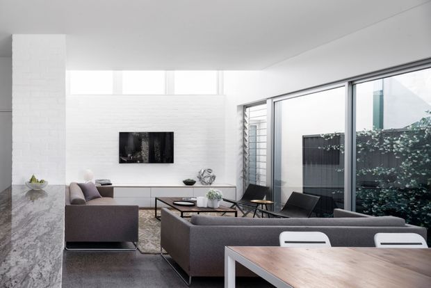 Bright contemporary open-plan living and dining area with white-painted brick walls, high clerestory windows, expansive aluminium-framed glazing, and a restrained grey and timber palette. A large sectional sofa faces a wall-mounted television, while a timber dining table occupies the foreground. Marble-look stone counters are visible at left, and a planted courtyard is seen through the glazing.