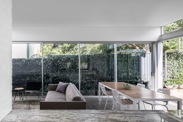 A bright, minimalist open-plan living and dining area with floor-to-ceiling glazing overlooking a planted courtyard. A neutral upholstered sofa faces the room, while a long natural timber dining table is paired with white chairs. The space has a smooth white ceiling, pale walls, dark-framed glazing, and a substantial grey stone kitchen island or benchtop in the foreground.