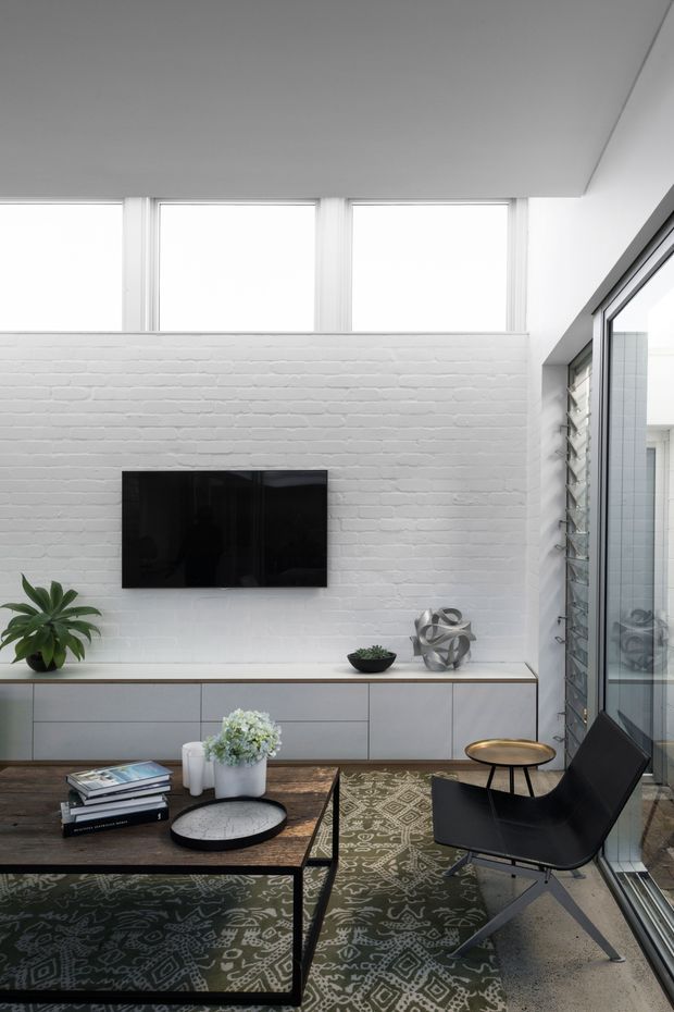 Bright contemporary living room with a white-painted exposed brick feature wall, wall-mounted television, low grey built-in cabinetry, high clerestory windows, and full-height glazed doors with louvred side panels. A dark timber coffee table, patterned rug, black lounge chair, plants, books, and sculptural decor furnish the space.