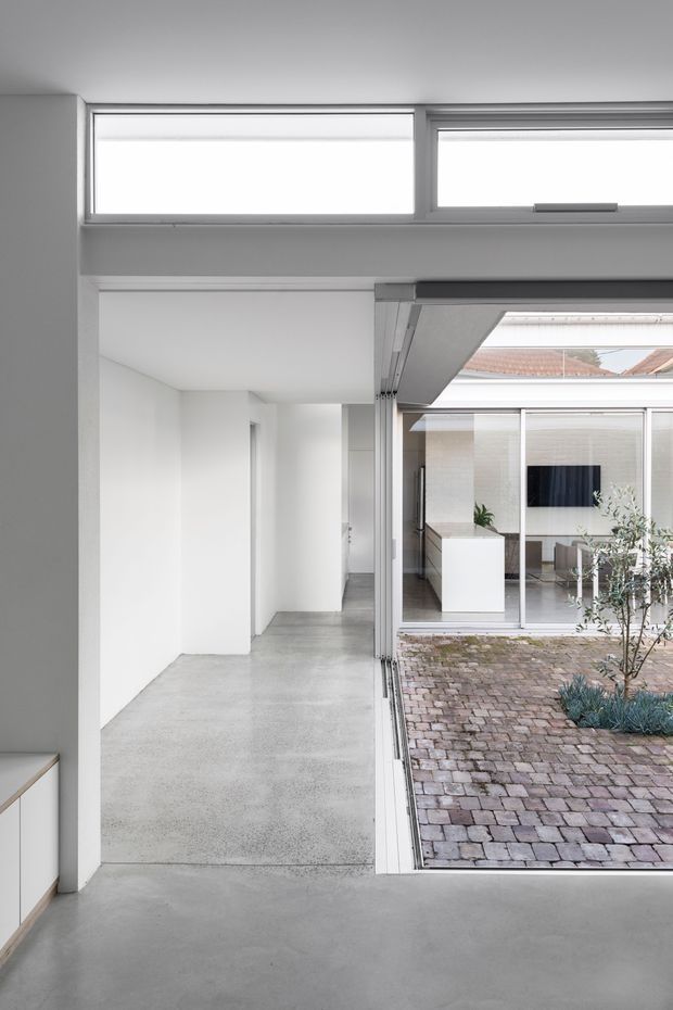 A bright minimalist interior overlooking a small landscaped courtyard. The open space has smooth pale concrete flooring, white walls, narrow clerestory windows, and large sliding glazed openings. A brick-paved courtyard with a small planted bed is visible outside, along with another white contemporary wing and a glimpse of a tiled roof. The image emphasises the indoor-outdoor connection and clean architectural geometry.