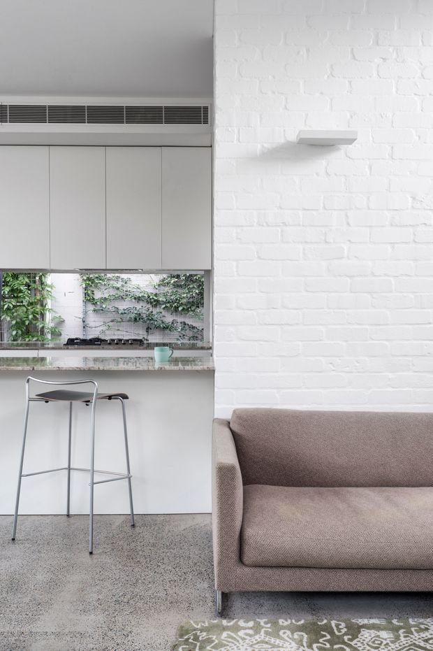 A bright, compact open-plan interior combining a minimalist kitchen and living area. The kitchen has flat-panel light cabinetry, a slim integrated ventilation grille, a pale stone or composite benchtop, and a gas cooktop overlooking a leafy courtyard. A simple metal bar stool sits at the peninsula. The adjacent living area features a taupe upholstered sofa against a white-painted exposed brick wall, with a small wall-mounted shelf and part of a patterned rug visible. The floor appears to be polished or speckled concrete/terrazzo.