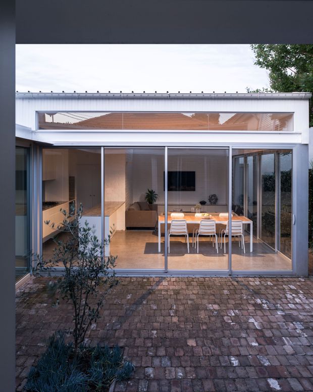 View from a paved courtyard into a light-filled contemporary house extension with full-height glazed sliding doors and a high clerestory window. The open-plan interior contains a compact kitchen with light timber cabinetry, a dining table with white chairs, and a small living area. The facade has a simple white rendered or painted finish, slim metal-framed glazing, and a low-profile roof edge.