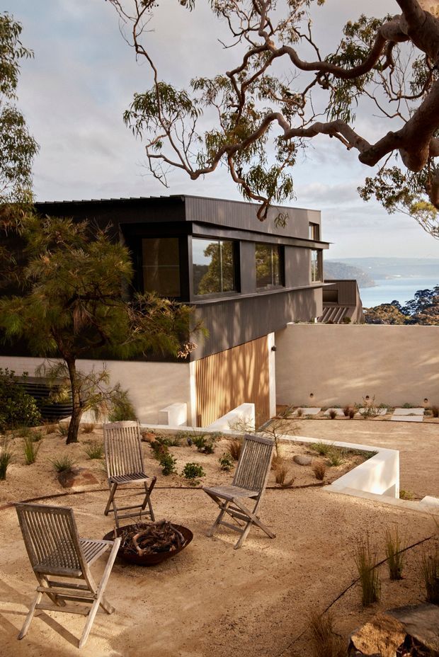 A modern coastal hillside residence viewed from a landscaped outdoor terrace. The dark corrugated-clad upper structure has extensive glazing and projects over warm rendered walls with timber screening. The drought-tolerant sandy landscape includes gravel planting beds, sculptural concrete edging, timber lounge chairs, and a fire bowl. A blue bay and distant shoreline are visible beyond the property, framed by native trees.