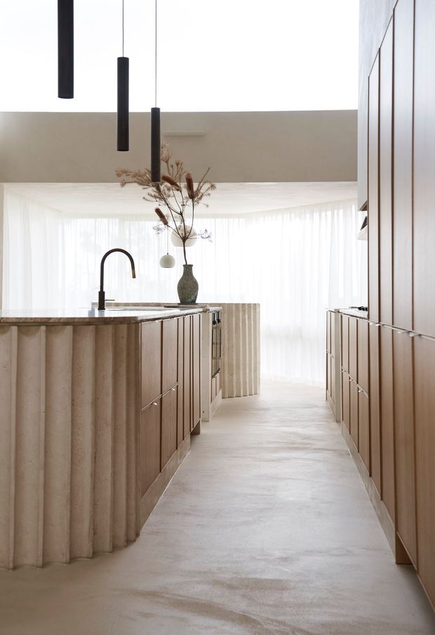 A refined contemporary kitchen viewed down a central aisle. The space features a sculptural fluted island with a dark stone benchtop, warm timber cabinetry along both sides, a black gooseneck tap, pale flooring, sheer full-height curtains, and minimalist black cylindrical pendant lights. A tall decorative vase and dried botanical arrangement sit near the rear of the kitchen, creating a calm, softly lit interior.