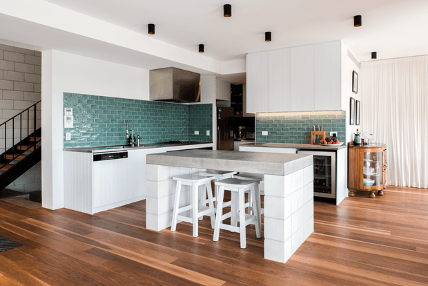 Bright open-plan kitchen with a large concrete-look island and white bar stools, white flat-panel cabinetry, and a glossy teal-green subway tile splashback. Warm timber flooring extends into the adjacent dining/living area, with exposed blockwork and a visible stairway at left. Black recessed or surface-mounted ceiling lights and warm wood accents add contrast.