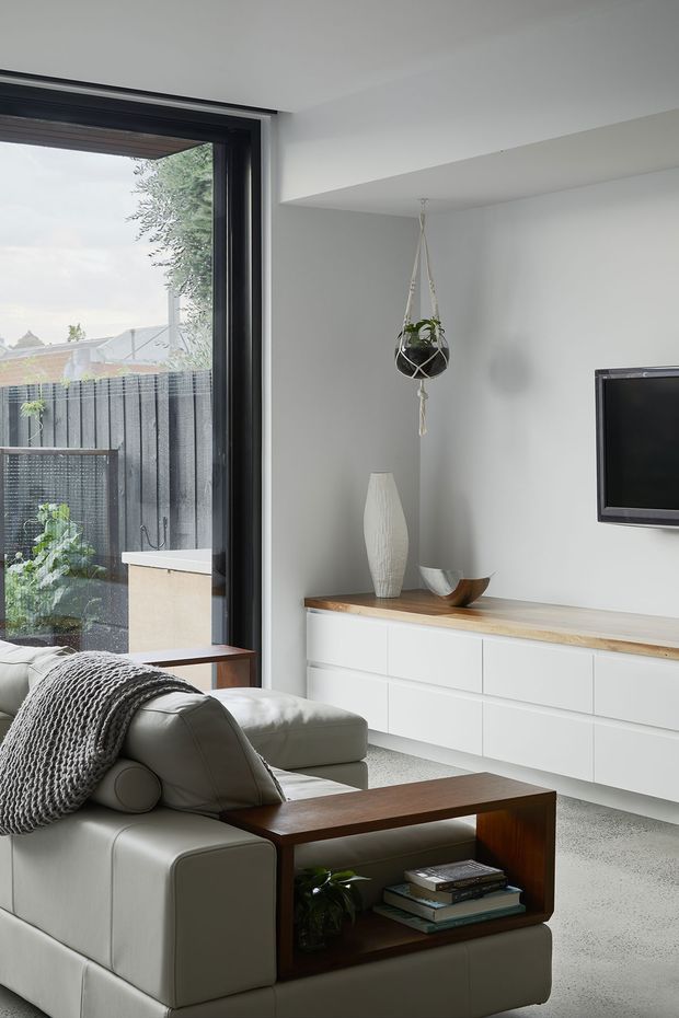 A bright, contemporary living room with a large black-framed glazed opening to an outdoor courtyard. The room features a pale upholstered sectional sofa, a dark timber open-sided coffee table, a white built-in media cabinet with a light timber benchtop, a wall-mounted television, and minimalist decorative objects. A hanging planter adds greenery near the window. The palette is predominantly white, light grey, and natural wood with black window framing.