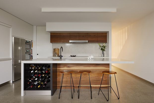A bright contemporary kitchen with a large white waterfall-edge island, integrated wine rack, timber-fronted cabinetry, stainless-steel appliances, and three slim bar stools. The room has polished concrete flooring, white walls and joinery, recessed cabinetry, and a simple linear ceiling light. Warm sunlight enters from the right, creating a clean yet inviting atmosphere.