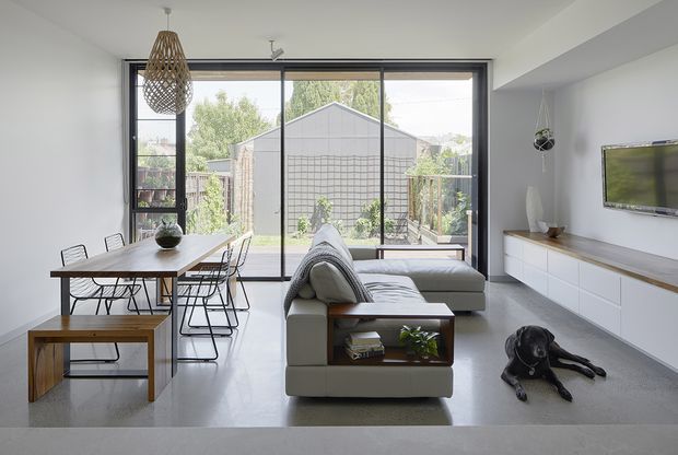 Bright open-plan living and dining area with a minimalist contemporary fit-out. A long timber dining table with black wire-frame chairs sits beside a low modular sofa, while a white built-in media cabinet spans the right wall beneath a wall-mounted television. Full-height black-framed glazing opens to a small outdoor area, and a woven pendant light adds texture.