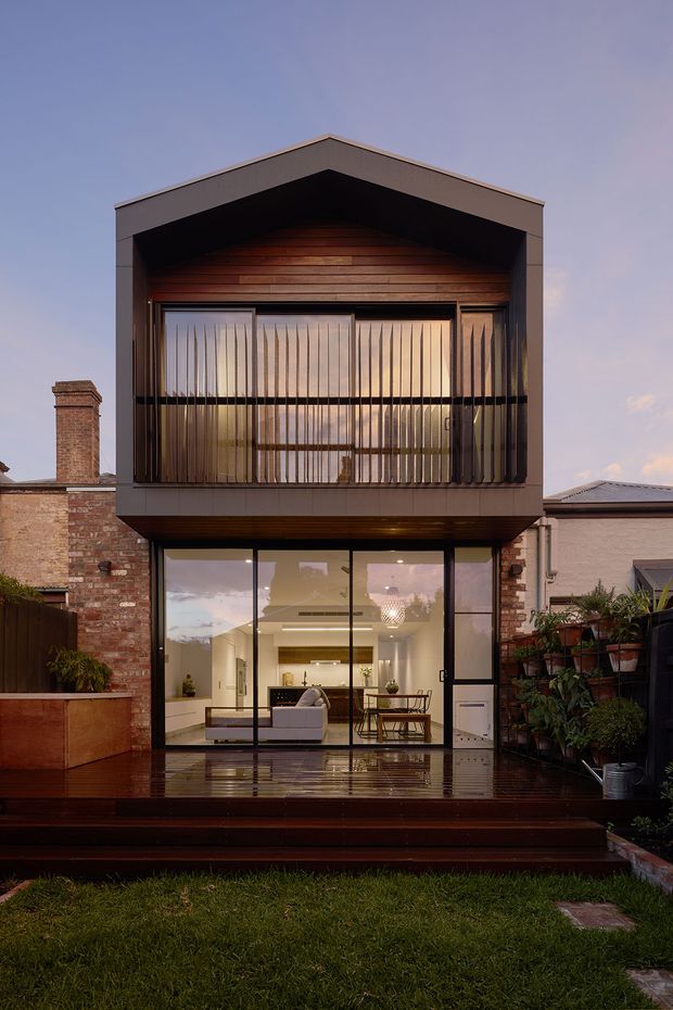 Rear exterior view of a compact two-storey residential extension with a prominent gabled upper volume. The upper level features dark framed glazing screened by vertical timber battens, while the ground floor opens through large glazed doors onto a wet timber deck. Existing brick walls and chimneys flank the contemporary addition, with a small landscaped backyard in the foreground.