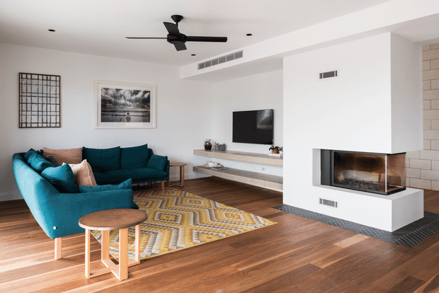 Bright contemporary living room with a deep teal sectional sofa, geometric patterned area rug, timber flooring, wall-mounted television and a minimalist built-in fireplace set within a white feature wall. A black ceiling fan, recessed lighting, framed artwork and floating media shelving complete the room.
