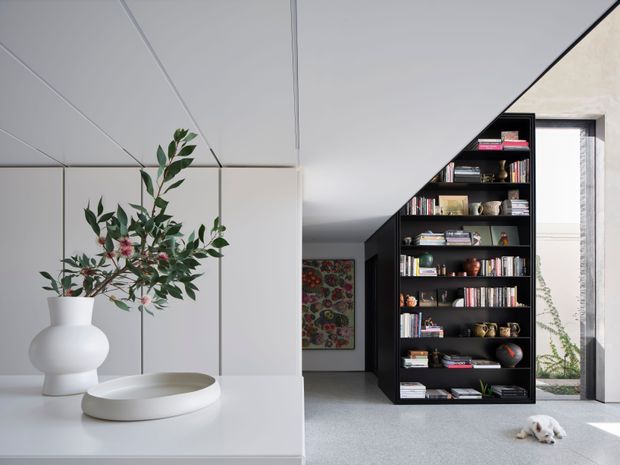 Bright contemporary interior featuring a white sculptural kitchen island or console in the foreground, a large matte white vase with leafy branches, and a dramatic full-height black built-in bookshelf forming a diagonal architectural partition. Books, ceramics, and decorative objects fill the shelving, with a narrow glazed opening to a planted courtyard beyond. Light grey flooring, white ceiling panels, and strong geometric lines create a refined, minimalist composition.
