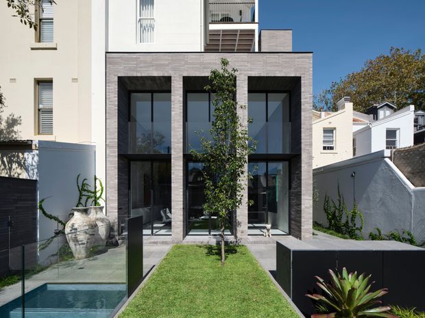 Contemporary multi-storey residential facade viewed from a landscaped front garden. The home features a dark grey masonry-clad central volume with tall black-framed glazing, flanked by lighter rendered neighbouring forms. A narrow lawn, glass-edged water feature or pool, black boundary walls, and mature planting define the foreground.