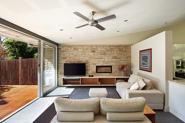 Bright contemporary living room with a large cream sectional sofa arranged around a timber media console and wall-mounted television. A linear fireplace is set into a textured exposed-stone feature wall, while full-height sliding glass doors open to a timber deck and fenced outdoor area. The room has a white ceiling with recessed lighting and a ceiling fan, dark tiled flooring, and a partial view into the kitchen.