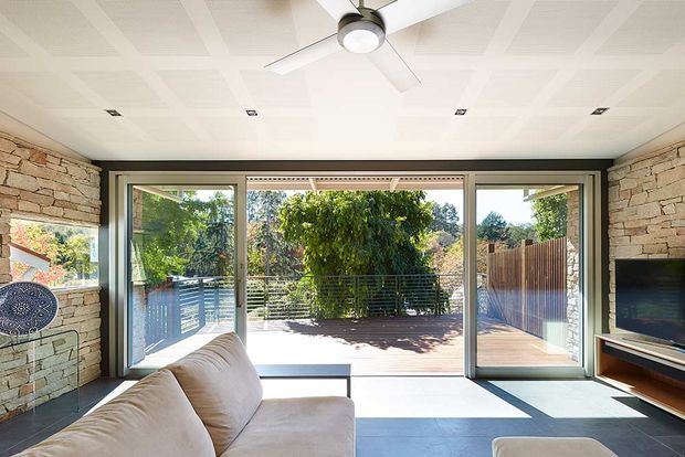 Bright contemporary living room opening onto a timber deck through large sliding glass doors. The room features exposed stacked-stone walls, a patterned white ceiling, dark-framed glazing, a ceiling fan, a neutral sectional sofa, and a wall-mounted television with low timber joinery. The outdoor deck and landscaped garden are visible beyond the glazing.