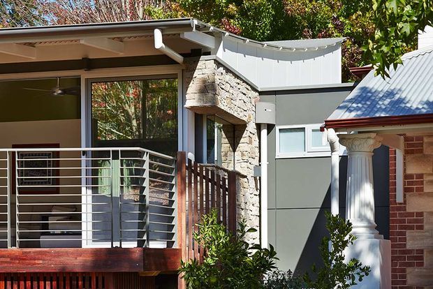 Exterior view of a compact residential extension combining a modern glazed living area with a balcony, grey rendered walls, a rough stone-clad projecting feature, and an adjoining traditional brick section with a classical column. Mature vegetation surrounds the building, and exposed roof drainage and string lighting are visible.