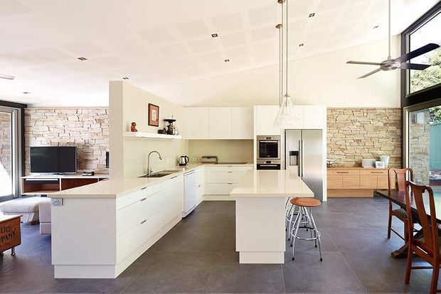Bright open-plan kitchen and living area with a large white island, integrated white cabinetry, stainless-steel appliances, and a dining zone. Pale exposed brick feature walls contrast with large dark grey floor tiles, while expansive black-framed glazing and a ceiling fan add a contemporary feel.