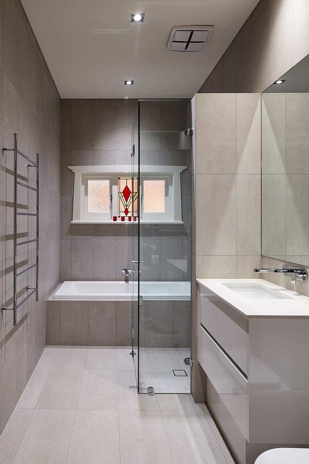 A narrow, contemporary bathroom viewed lengthwise, featuring full-height grey-beige tiled walls and matching large-format floor tiles. A clear frameless glass shower enclosure occupies the center, with a built-in bathtub beneath a frosted-glass window and decorative stained-glass panel. On the right is a white wall-mounted vanity with integrated basin, mirrored wall cabinet or splashback, and chrome tapware. A chrome heated towel rail is mounted on the left wall, with recessed ceiling spotlights and a concealed air-conditioning vent.