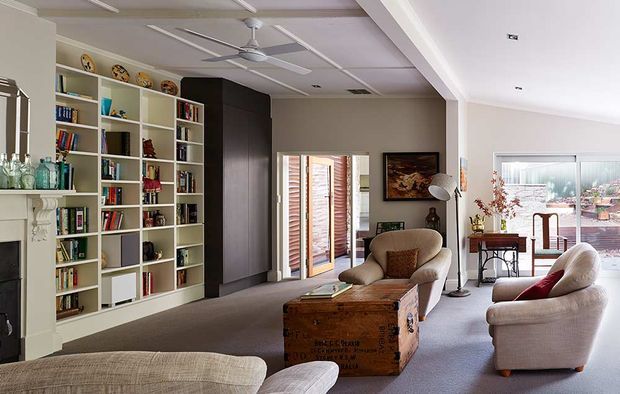 Spacious, bright living room with a neutral palette, carpeted floor, a large built-in white bookcase filled with books and objects, dark timber cabinetry, a decorative fireplace surround, upholstered armchairs and sofas, and a rustic wooden trunk used as a coffee table. A ceiling fan, framed artwork, console table, and large glazed openings complete the relaxed traditional interior.