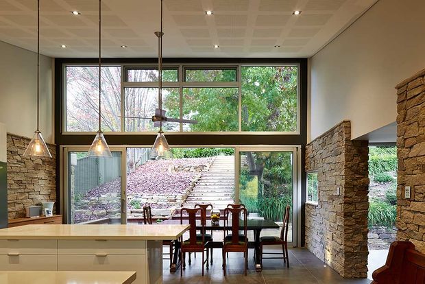 Open-plan interior showing a kitchen island in the foreground and a dining area beyond, with large timber-framed windows, pendant lights, exposed stone walls and views to a landscaped garden. The room has a warm, earthy palette and strong indoor-outdoor connection.