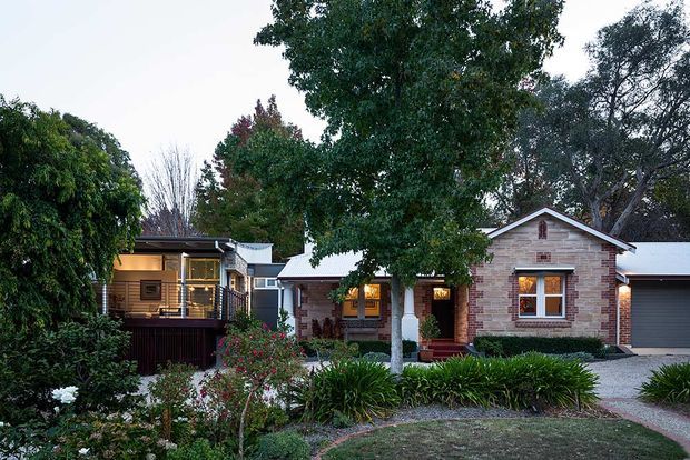 Single-storey residential home viewed from the landscaped front garden, featuring a brick facade, pitched gabled roofs with light metal roofing, timber-framed windows and doors, a covered entry, and an integrated garage. Mature planting, garden beds, and a gravel driveway surround the house.