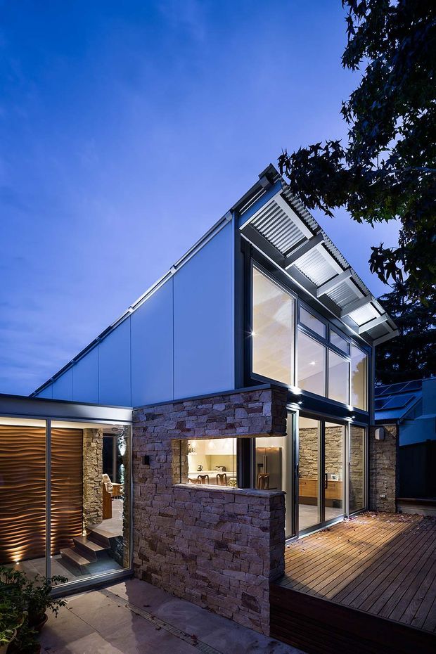 Twilight exterior view of a contemporary multi-level residence organized around a timber deck courtyard. The building combines dark metal framing, large glazed openings, pale blue-grey wall panels, and prominent rough-stacked natural stone walls. A deep projecting roof canopy with slatted underside shades the upper glazing, while a sculptural timber entry screen and illuminated interiors add warmth.