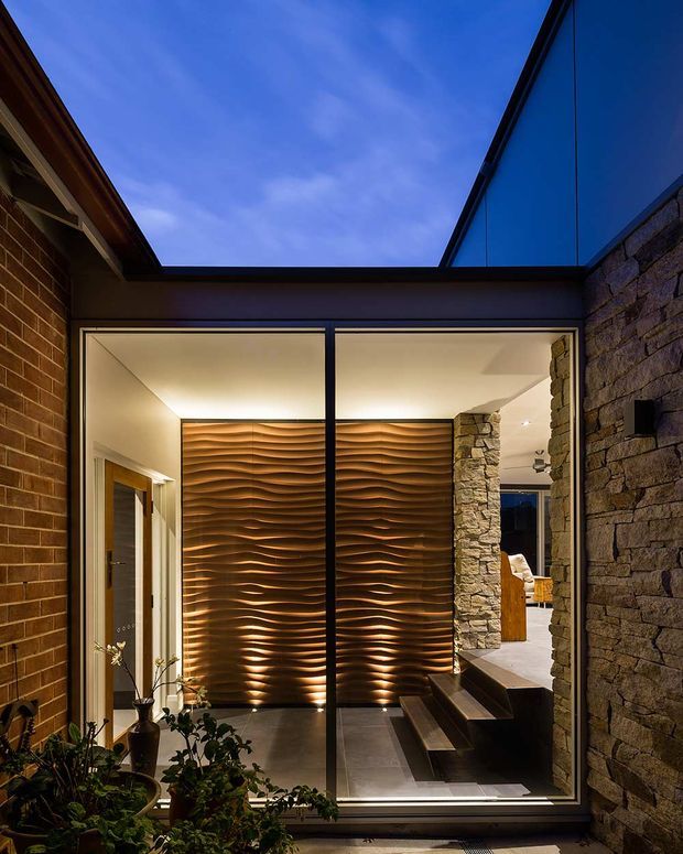 A dramatic contemporary residential entry courtyard viewed from outside, framed by dark brick and rough natural-stone walls. A large black-framed glazed opening reveals a warmly lit foyer with a sculptural rippled timber feature wall, indoor planting, tiled flooring, and stepped circulation. The open-to-sky void and strong warm/cool contrast create a refined, architectural atmosphere.
