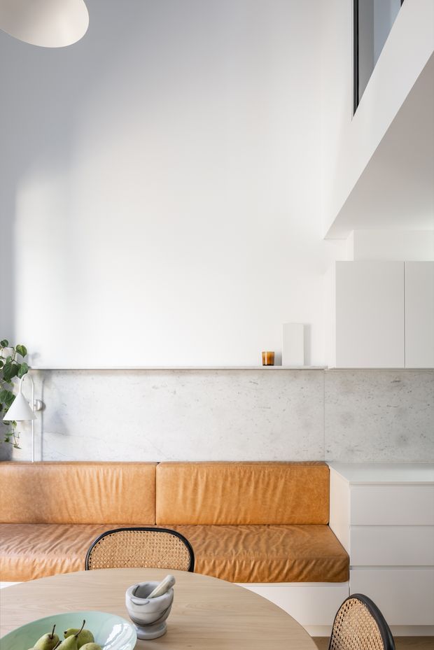 Bright minimalist open-plan living and dining area with a tan leather built-in sofa beneath a pale concrete or stone ledge, white walls and fixed cabinetry, and a light timber dining table with cane chairs. A tall ceiling, clerestory-style glazing, simple pendant, and restrained decor create a clean contemporary interior.