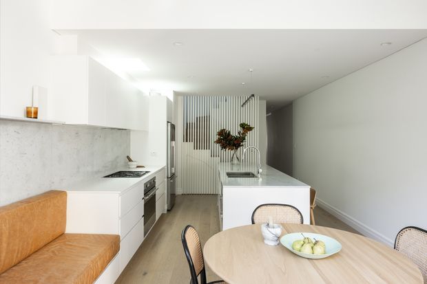 Bright, compact open-plan kitchen and dining area with a warm tan built-in bench seat, white cabinetry, pale stone or concrete-look splashback, central island with integrated sink, light timber-look flooring, and a round light-wood dining table with cane-backed chairs. A vertical slatted screen and floral arrangement add texture at the rear, while recessed ceiling lighting and clean white surfaces create a contemporary, minimalist feel.