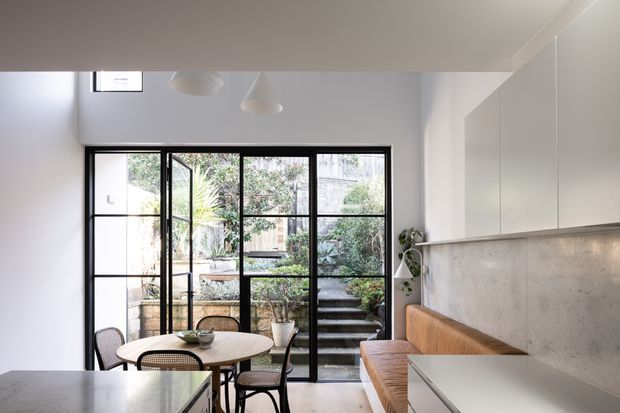 Bright contemporary kitchen-dining area with white flat-panel cabinetry, a concrete-look splashback, pale stone island benchtop, and a built-in timber bench seat. Black-framed floor-to-ceiling glazing and a glazed door open onto a planted courtyard with steps. A round dining table and woven chairs sit beside the window, while simple pendant lights hang above.