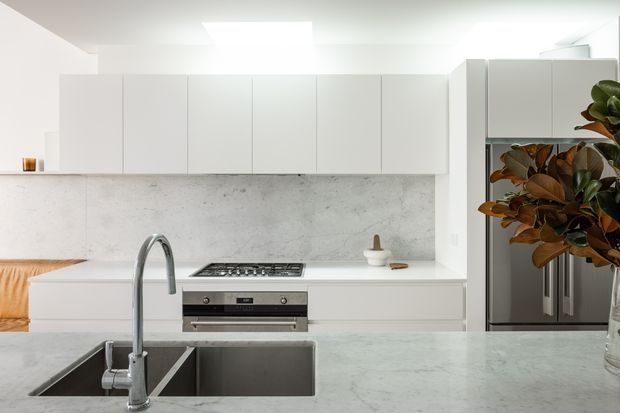 Bright, minimalist contemporary kitchen with extensive matte white handleless cabinetry, a pale concrete-look splashback and island benchtop, integrated stainless-steel cooking appliances, and a large double-bowl sink with a curved chrome tap. A stainless-steel refrigerator and indoor plant are visible at the right, while a small glimpse of adjacent seating appears at the left.