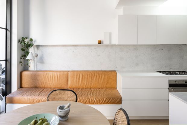 Bright open-plan kitchen and dining area with a built-in tan leather banquette, round light-wood dining table, woven cane chairs, white flat-panel cabinetry, a pale concrete-look splashback, and black-framed glazing. The space has a clean contemporary fit-out with warm natural accents.