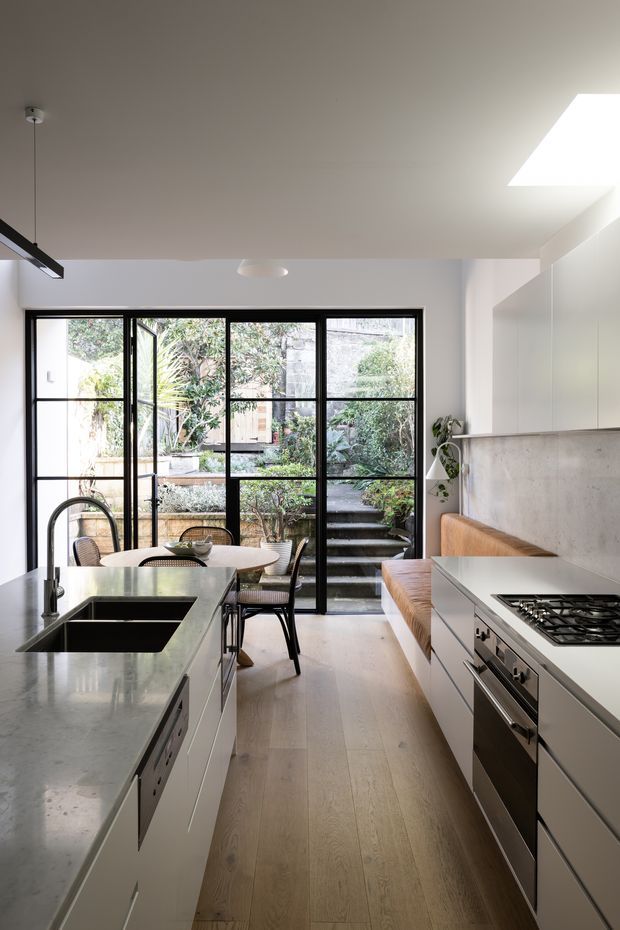 Bright contemporary kitchen with a large central island, integrated double sink, pale stone benchtops, white built-in cabinetry, stainless-steel appliances, and a warm timber floor. Black-framed glazed doors open to a planted courtyard with steps, while a built-in upholstered bench and dining table occupy the rear of the room. A skylight and pendant lighting provide additional illumination.