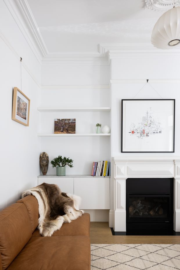 Bright, refined living room with white walls, ornate ceiling cornices and a classic white mantel fireplace. Built-in white shelving and cabinetry display books, plants, ceramics and framed artwork. A warm tan leather sofa with a fur throw occupies the foreground, complemented by a light geometric rug and a sculptural pendant light.