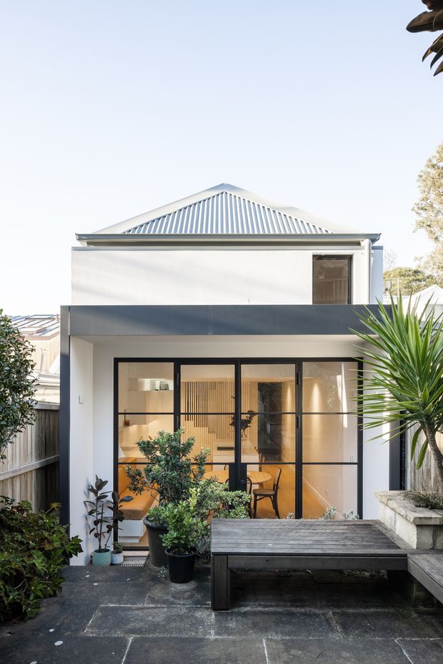 A bright rear exterior view of a compact two-storey home extension or renovation. The white rendered facade has a traditional pitched metal roof with gable detailing, a dark horizontal balcony or fascia element, and large black-framed glazed doors opening onto a paved courtyard. Potted plants, raised masonry garden beds, and a timber outdoor bench or table occupy the foreground.