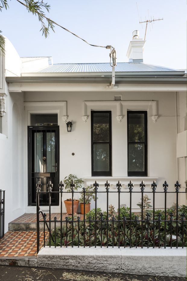 Street-facing heritage-style townhouse facade with a white rendered exterior, decorative moulded window surrounds, black-framed windows and glazed front door, ornate black metal fencing, a small planted frontage, and a patterned tiled entry path. The low-pitched corrugated metal roof and prominent classical chimney add character to the compact urban frontage.