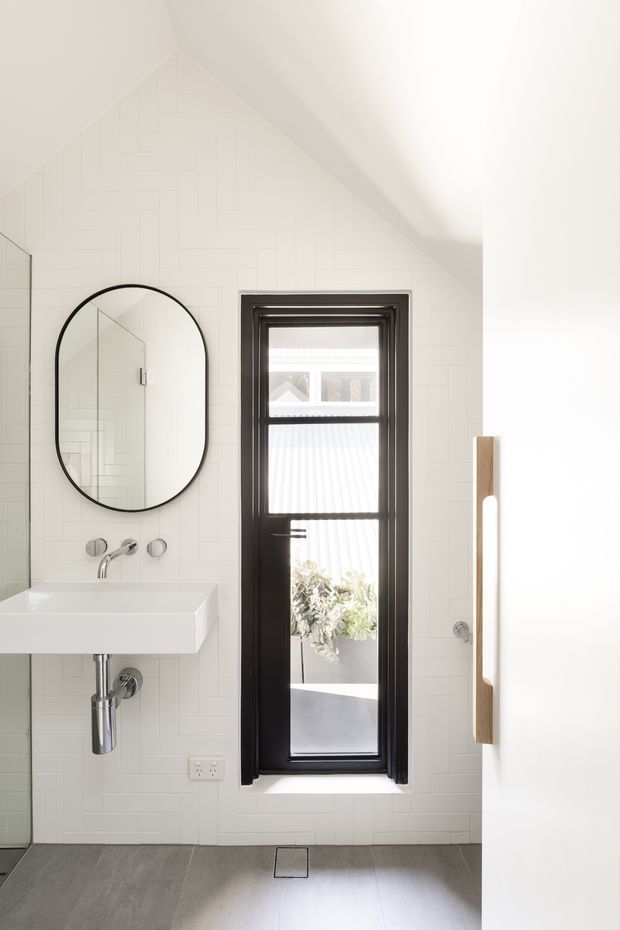 Bright, compact contemporary bathroom with a high sloping ceiling and white tiled walls. The room features a wall-mounted white rectangular basin with brushed metal tapware, a large black-framed oval mirror, a narrow black-framed window, grey floor tiles, and a vertical timber feature element at right. Natural light creates an airy, clean atmosphere.