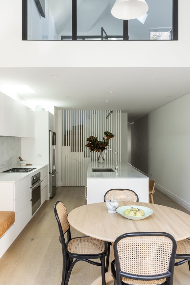 Bright contemporary open-plan kitchen and dining area with white cabinetry, a pale stone island benchtop, integrated appliances, light timber flooring, and a round timber dining table with woven cane chairs. A vertical slatted screen separates the kitchen from the hallway, while black-framed upper-level glazing and a large white pendant add architectural interest.