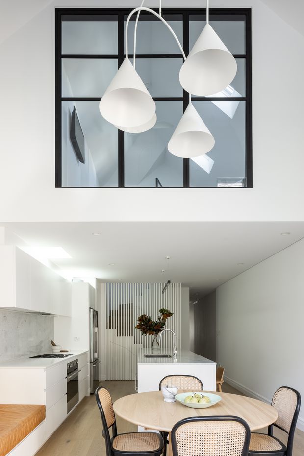 Bright contemporary open-plan kitchen and dining area with white cabinetry, pale timber flooring, a light stone island benchtop, and a round timber dining table with cane-backed chairs. A black-framed internal window or glazed partition sits above the kitchen, with three sculptural white pendant lights suspended in front of it. Vertical slatted screening and floral branches add a subtle decorative feature.