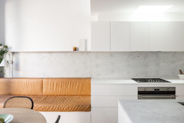 Bright, compact open-plan kitchen and dining area with a built-in tan leather banquette along the rear wall, white cabinetry, a pale mottled concrete-look splashback, and a white marble-look island benchtop. A gas cooktop and built-in oven are integrated into the minimalist kitchen joinery, with a small round dining table and cane-backed chair in the foreground. The space has clean contemporary lines and abundant diffuse light from a roof opening or skylight.
