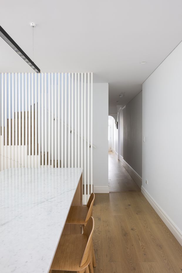 Bright minimalist interior view from a kitchen or dining area toward a long hallway. A large white marble-look island or dining counter with timber stools occupies the foreground, while a white vertical timber slat screen partially separates the space from a stairwell. The hallway has pale grey walls, white trim, recessed lighting, and warm natural timber flooring. A slim black linear pendant light runs above the counter.