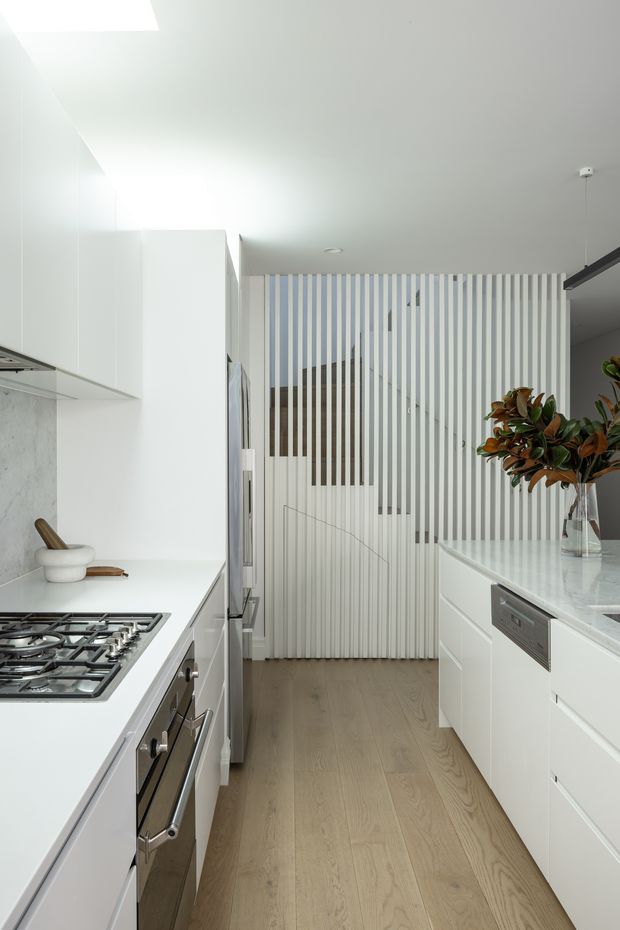 A bright, narrow contemporary kitchen with white full-height cabinetry, a gas cooktop and built-in oven along the left side, and a white island with integrated dishwasher on the right. A pale stone-look splashback, light timber flooring, and a vertical white slatted screen partially reveal a staircase at the rear. A vase of leafy branches sits on the island, adding a natural accent.