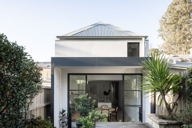 Front exterior view of a compact two-storey urban residence with a light rendered facade, dark horizontal balcony or awning band, large black-framed glazed doors, and a corrugated metal hipped roof. The home is framed by a small landscaped courtyard with established plants and a paved entry area.