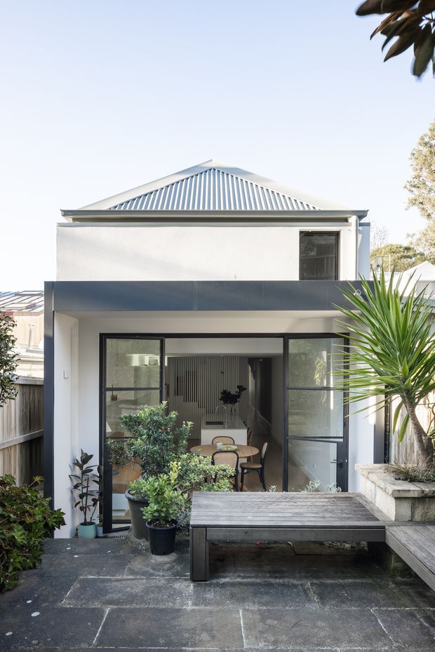 A contemporary two-storey residential extension viewed from a paved rear patio. The facade combines smooth white rendered walls, dark metal-framed glazing, a projecting dark horizontal balcony or awning band, and a corrugated metal gable roof with visible vertical ribbing. Large glazed doors open onto a compact courtyard with timber outdoor bench seating, raised planters, and potted greenery. A dining area and interior hallway are visible through the glazing.