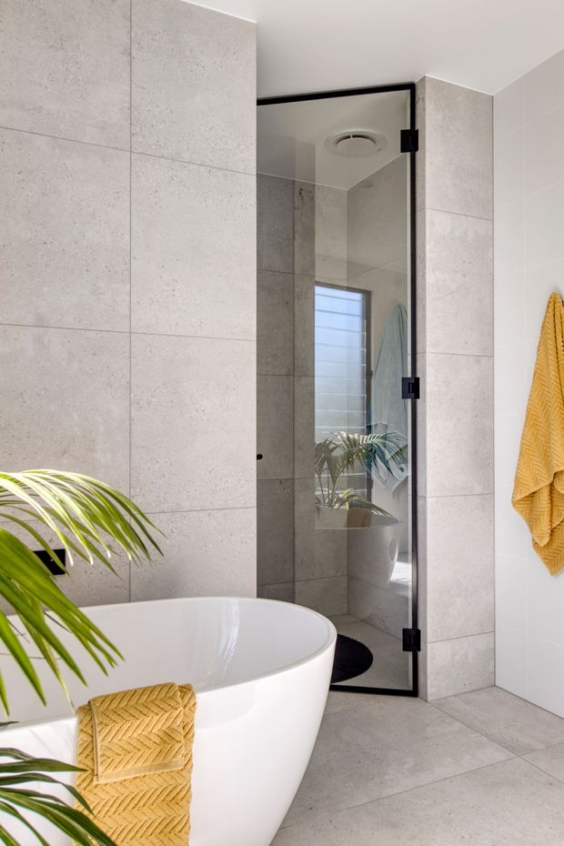 Bright contemporary bathroom featuring a freestanding white oval bathtub, large light-grey concrete-look wall and floor tiles, and a frameless glass shower enclosure with black hardware. A small window, potted tropical plant, and mustard-yellow textured towels add natural and warm accents.