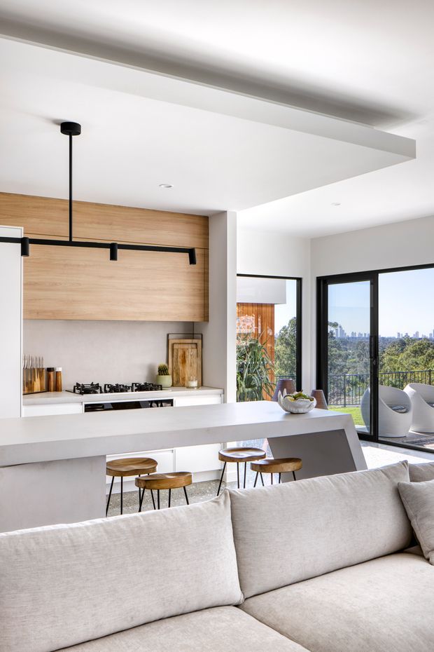A bright, open-plan contemporary interior combining a kitchen, dining island and living area. The kitchen features light timber wall cabinetry, white cabinetry and benchtops, black linear pendant lighting, and integrated black-framed appliances. A large white island with timber stools separates the kitchen from a light neutral sectional sofa in the foreground. Black-framed floor-to-ceiling sliding doors open to a balcony or terrace with leafy views and a distant urban skyline.