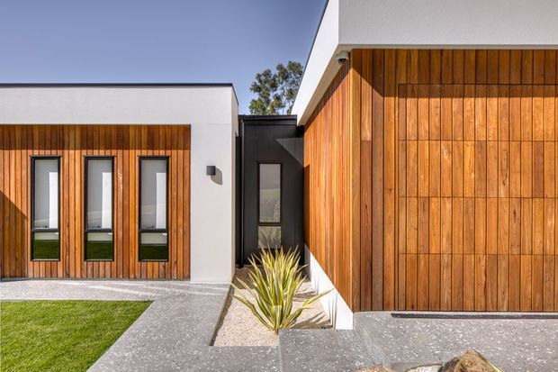 Exterior view of a contemporary single-storey residential building with strong geometric volumes, warm vertical timber wall cladding, white rendered sections, and a dark recessed entry. Narrow vertical windows punctuate the timber facade, while concrete paving, gravel, and sculptural drought-tolerant planting define the front approach.