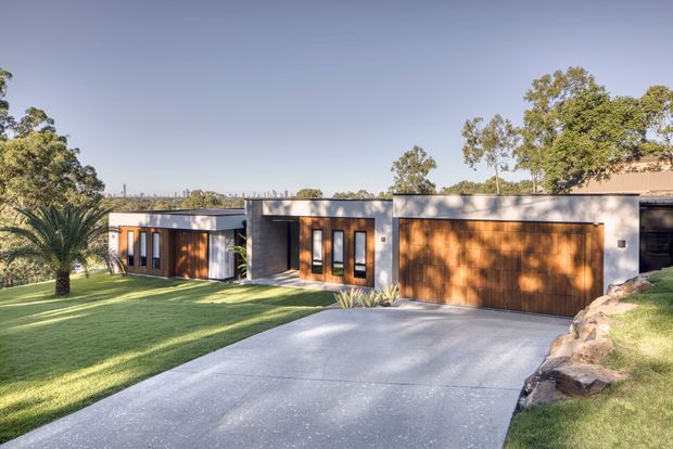 A low-slung contemporary residence viewed from the driveway, with a flat-roofed white and dark-grey facade, extensive warm timber-clad garage and entry elements, tall narrow windows, and a broad concrete driveway. The house sits within a landscaped grassy site with mature trees and rock edging.