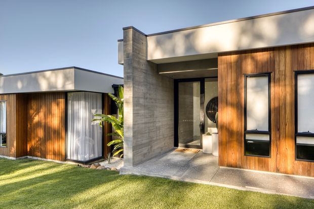 A contemporary single-storey residential facade viewed from the landscaped front lawn. The home features a flat roof, warm vertical timber cladding, large dark-framed windows, a recessed entry, and a prominent exposed concrete wall and porch surface. Strong late-afternoon light creates dappled shadows across the facade.